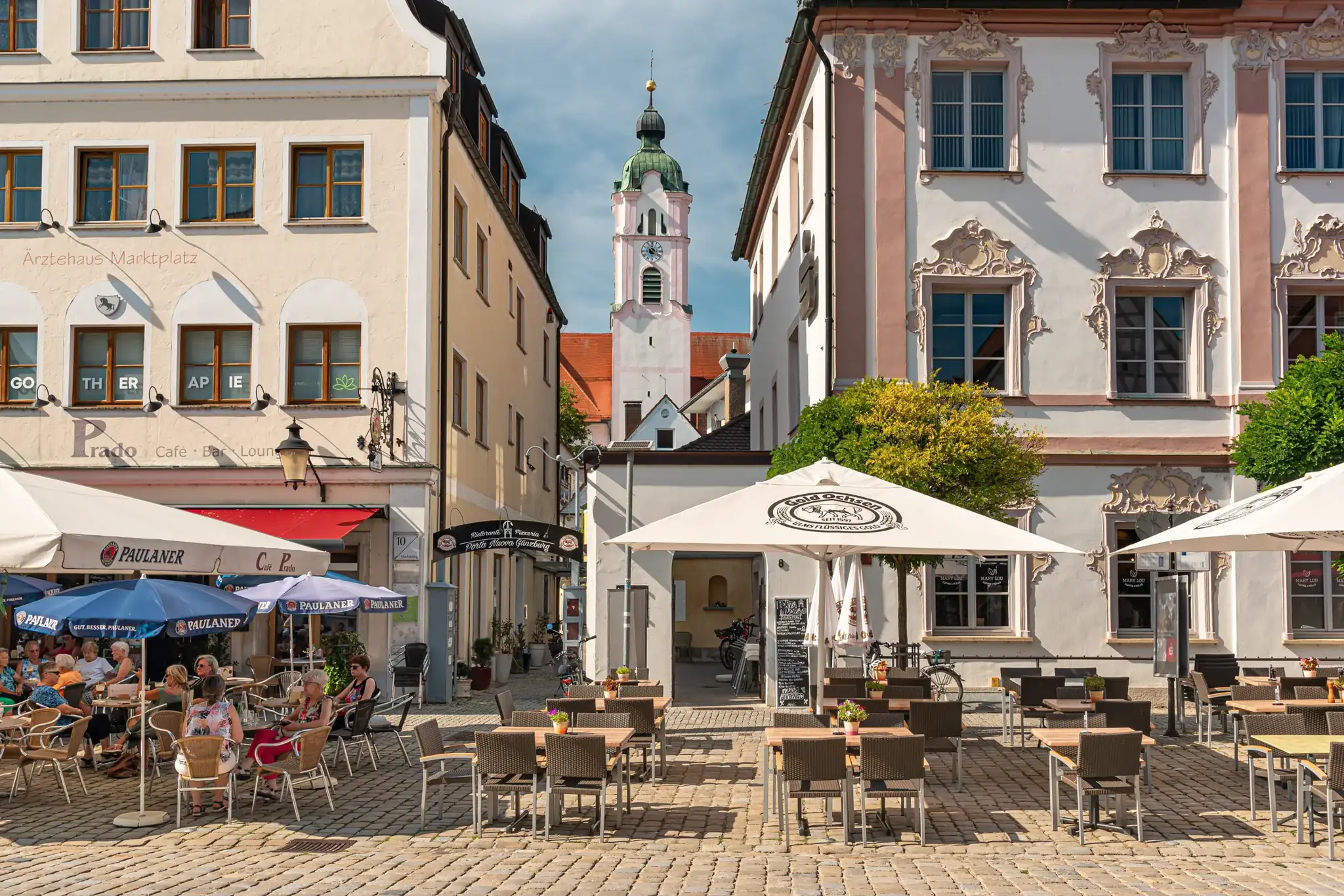 Marktplatz in Günzburg bei Sonne, Cafés vor barocken Häusern, dahinter der Turm der Frauenkirche