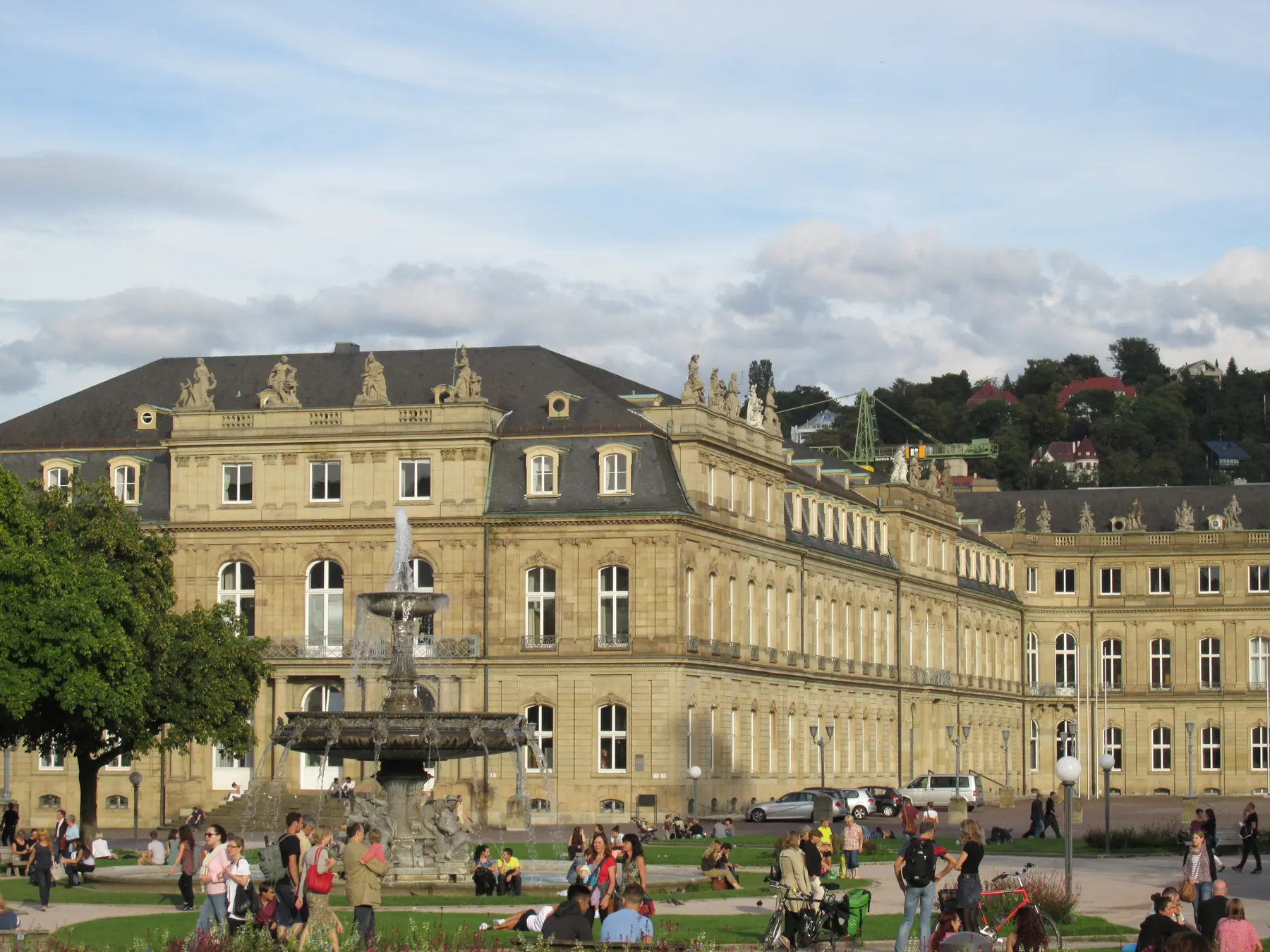 Stuttgarter Schlossplatz mit Brunnen, Neuem Schloss und Menschen auf der Wiese, dahinter die Hänge der Stadt