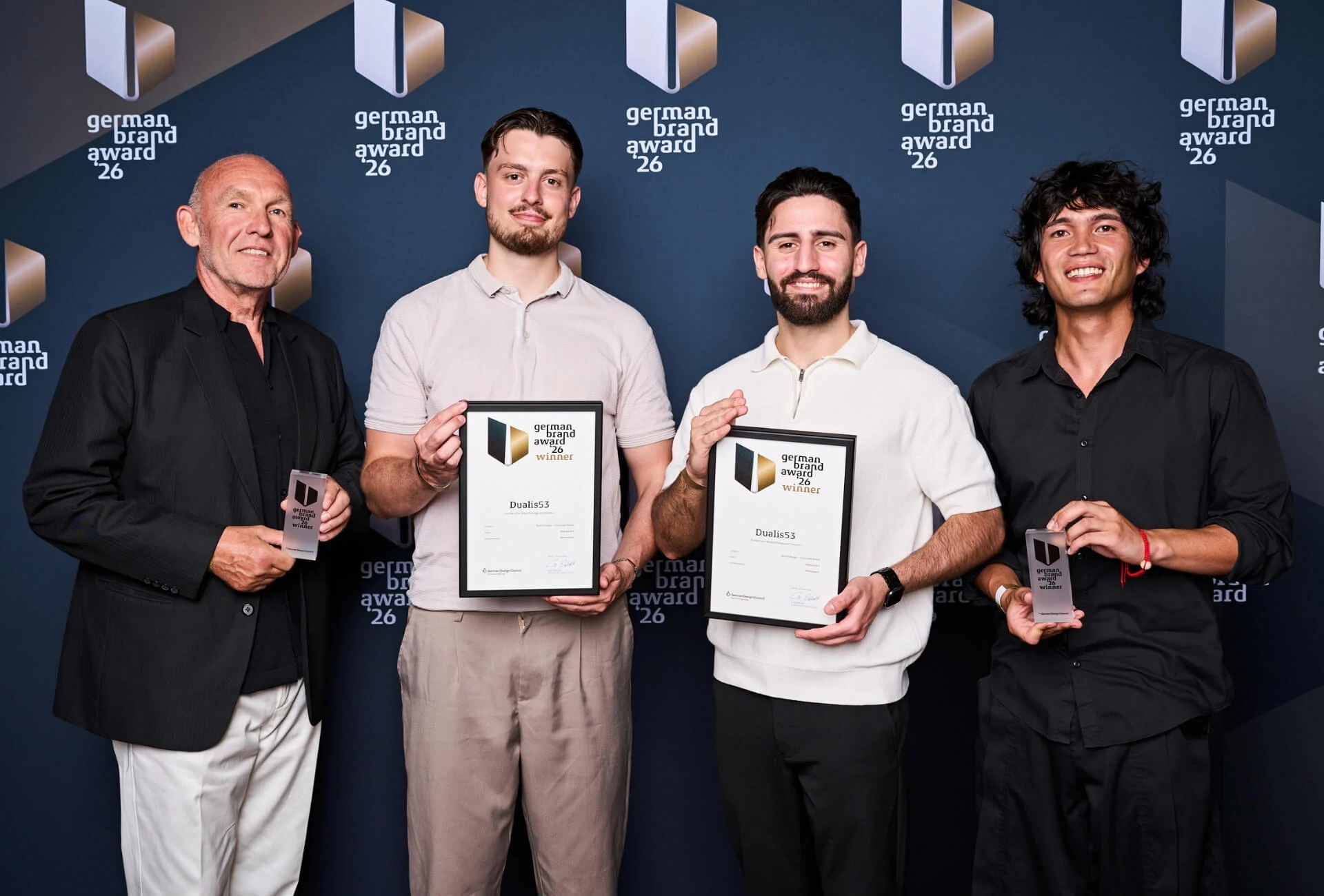 Vier Beteiligte mit Urkunden und Trophäen für Dualis53 vor der Fotowand des German Brand Award 2026